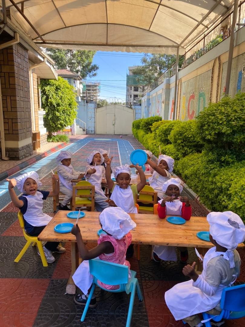 Young students in chef hats sitting around an outdoor table during a hands-on play-based learning activity